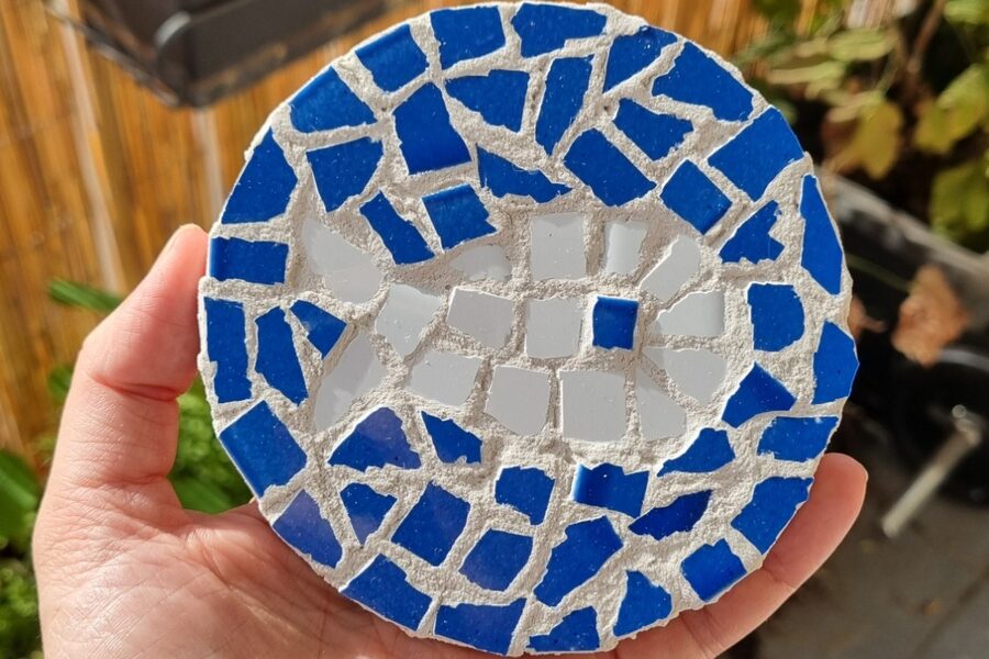 Dessous de plat rond en mosaïque, tenu dans la main : tesselles de carrelage bleu tout autour et blanches au centre, prises dans un joint clair