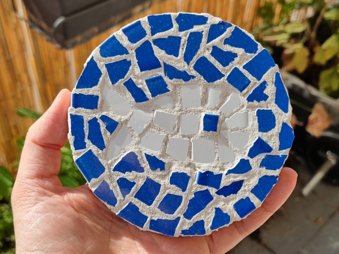 Dessous de plat rond en mosaïque, tenu dans la main : tesselles de carrelage bleu tout autour et blanches au centre, prises dans un joint clair