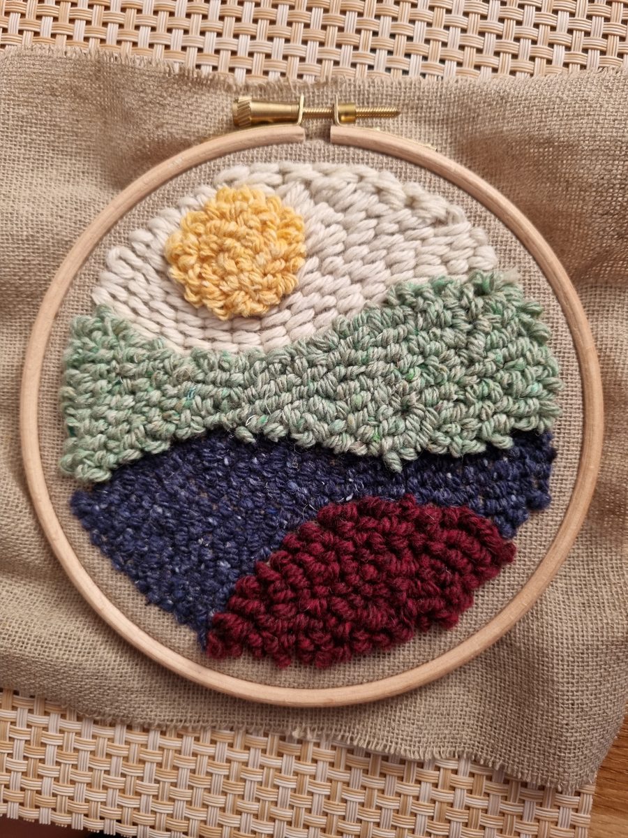 Paysage brodé au punch needle sur tambour en bois : soleil jaune, collines vertes, lac bleu et colline bordeaux, les boucles de laine en relief bien visibles