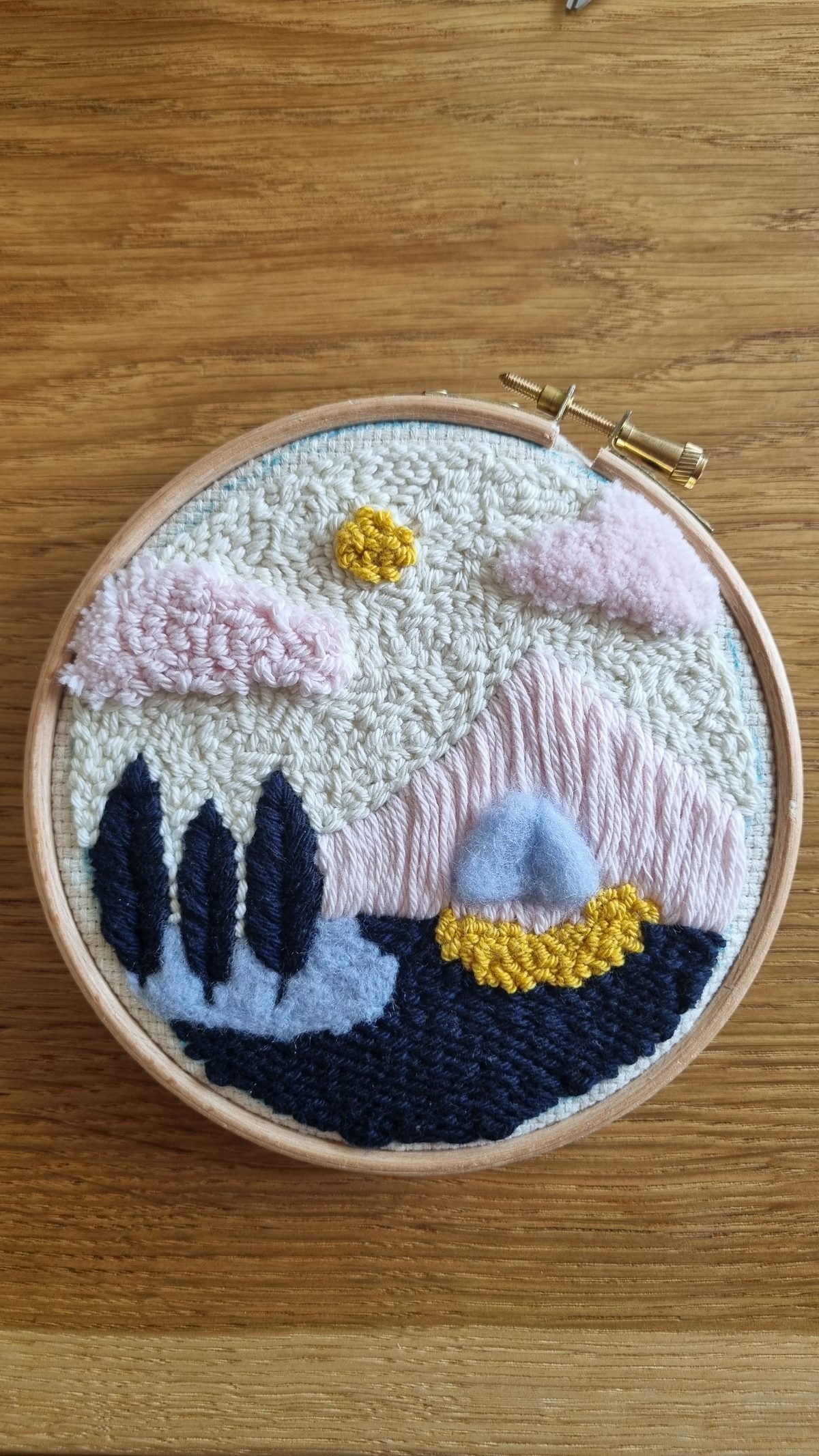 Paysage combinant punch needle et feutrage à l'aiguille : nuages roses en relief, soleil jaune, montagne rose et lac bleu marine sur tambour